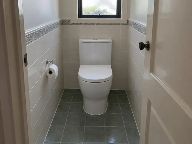 White toilet in a small tiled bathroom with a window and decorative border in Minmi
